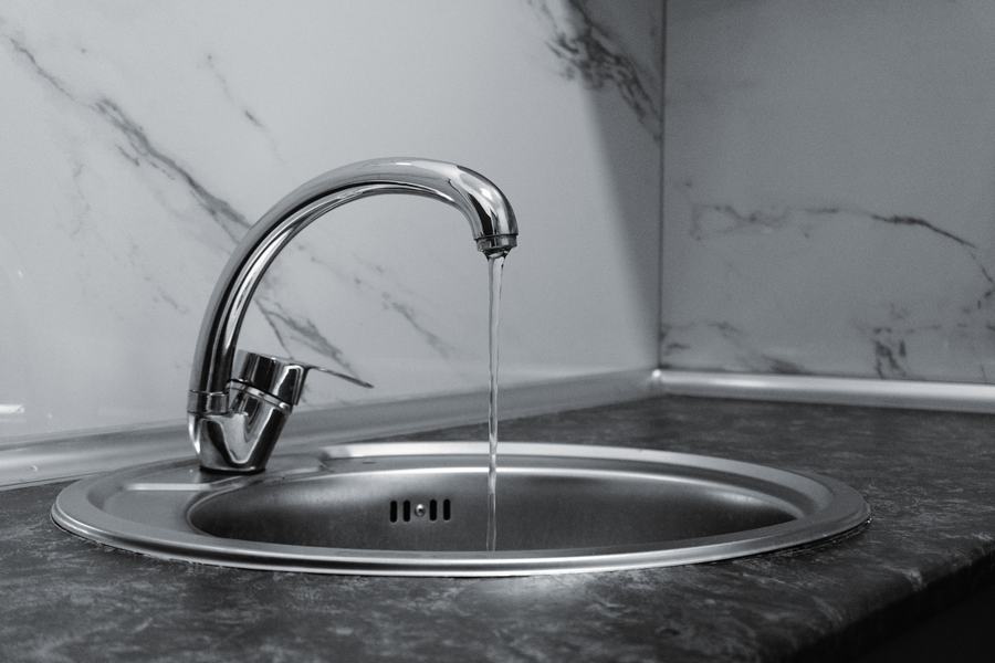 Robinet chromé au bec recourbé avec un filet d'eau qui coule dans un évier en inox, devant un mur en marbre